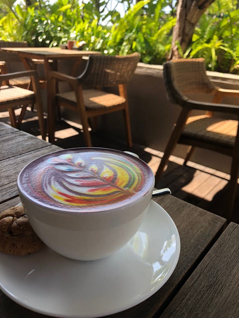 Vivid rainbow latte art in a white cup with a cookie on an outdoor wooden table.