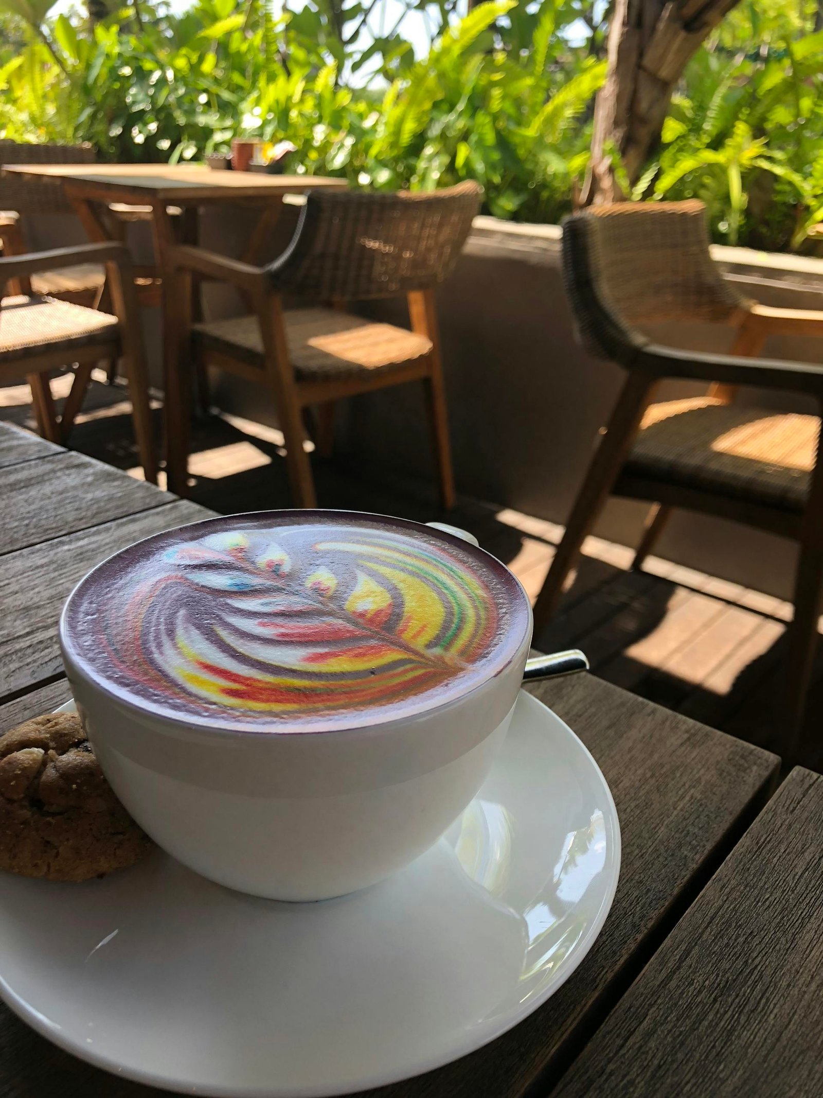Vivid rainbow latte art in a white cup with a cookie on an outdoor wooden table.